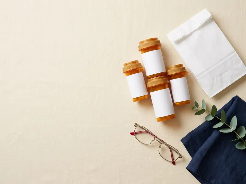 Four prescription bottles, a folded pharmacy bag and reading glasses on a table.